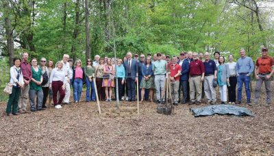 A group of current and retired faculty, and staff gather behind a newly planted tree, shovels, and a tarp among a mulched forested area  
