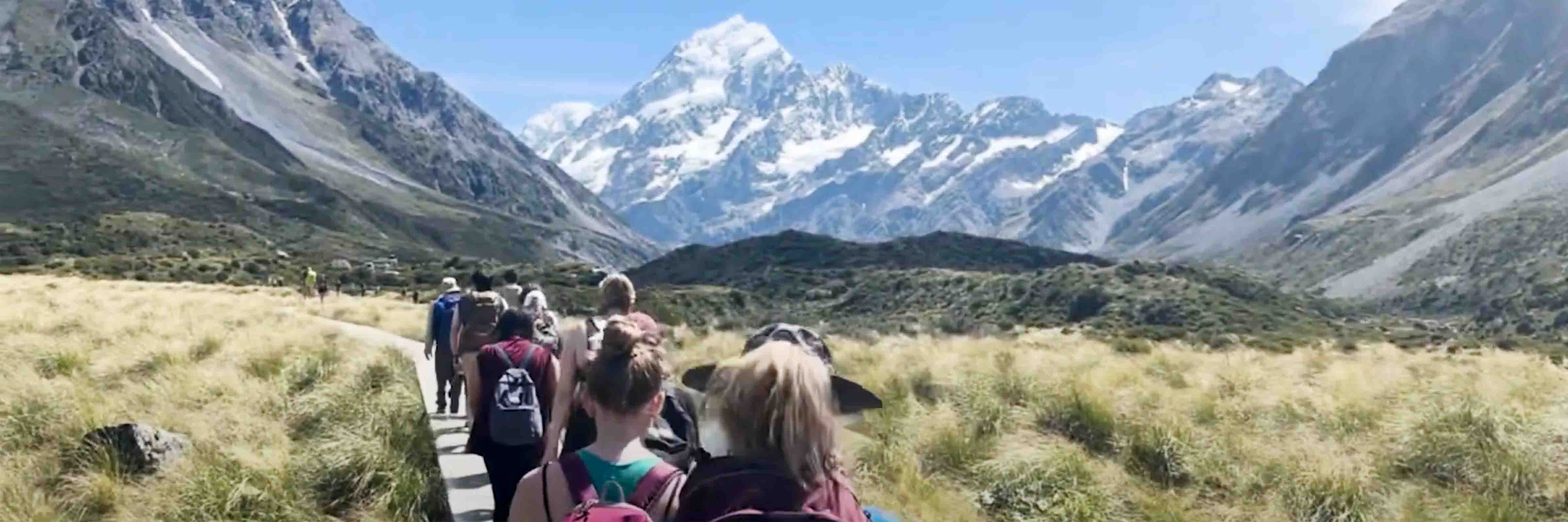 Students walking on a raised wooden path over grassland with snow capped mountains in the distance