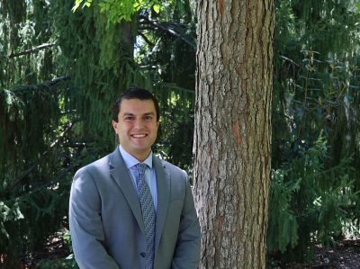 Man with dark brown hair in a gray suit with a blue tie outdoors in front of a tree