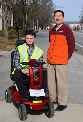 Student on a mapping scooter stand next to man with short hair in a maroon and orange jacket.